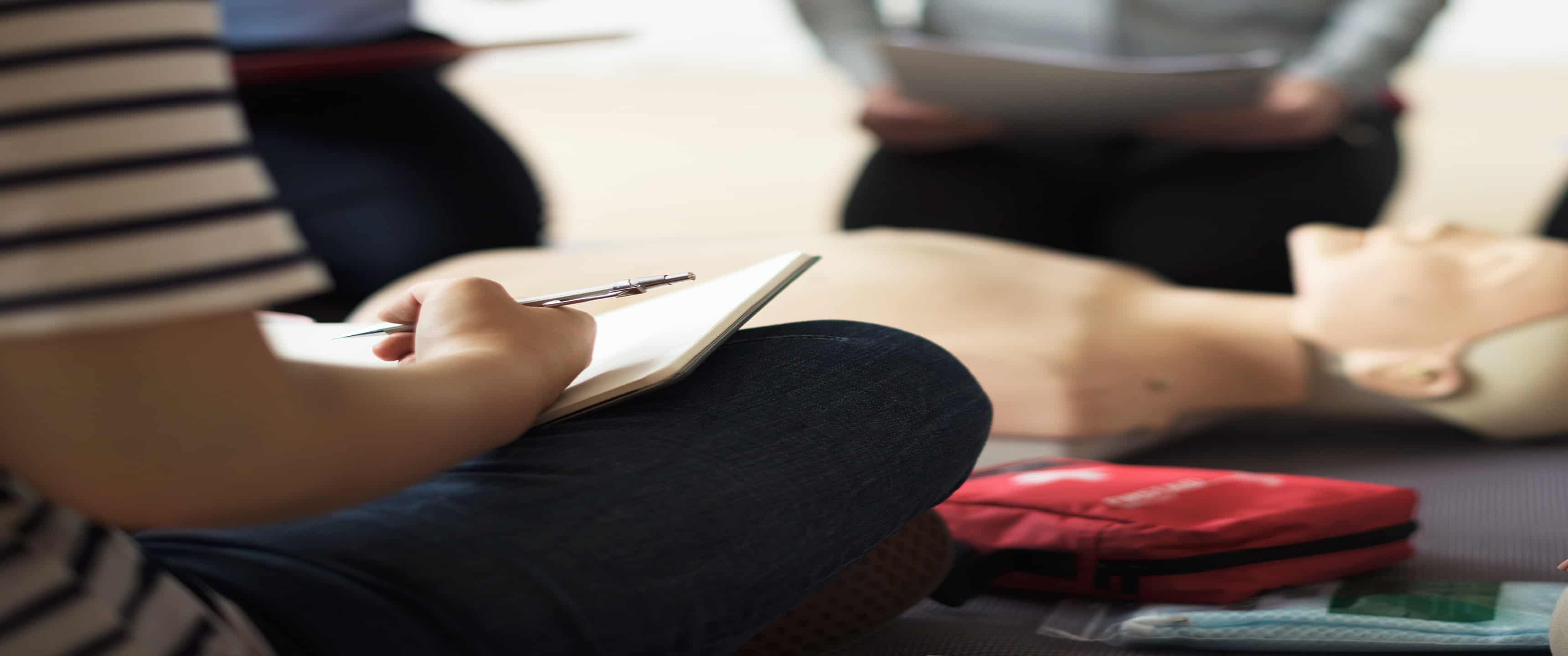 a person sitting on the floor with legs crossed, a notebook, a pen, with other people surrounding a CPR mannequin